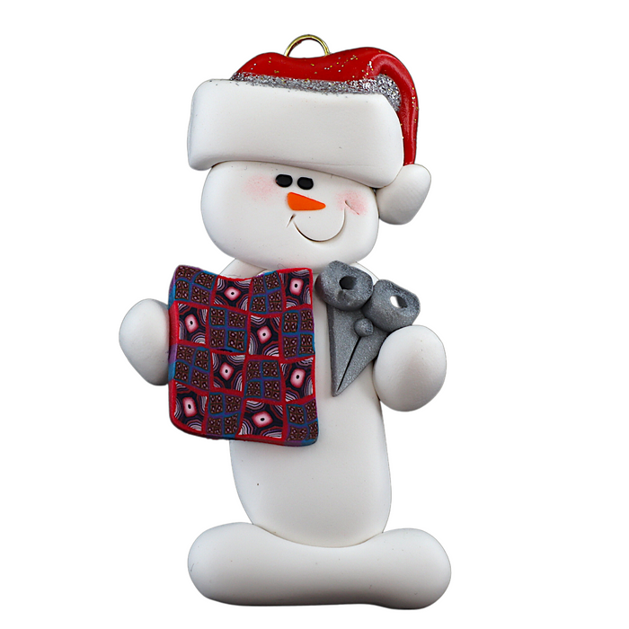 Ornament of white snowman in red hat holding a quilt block and pair of scissors.