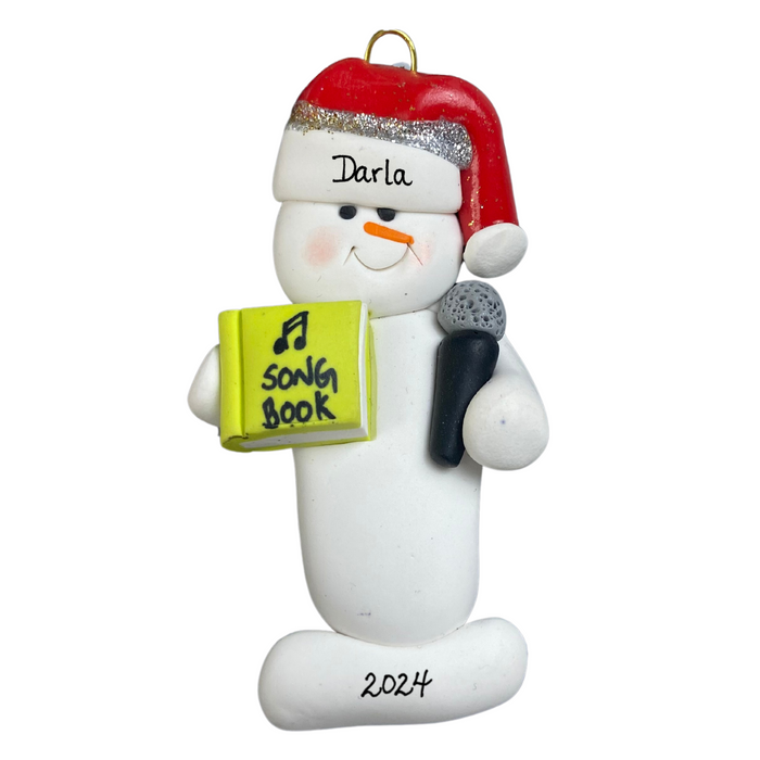 Ornament of white snowman in red hat holding a microphone and green music book, personalized with name.