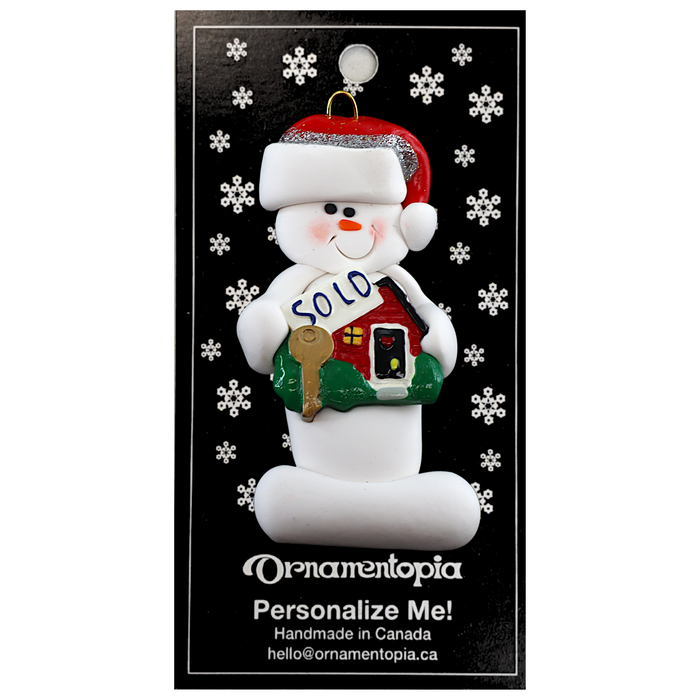 Ornament of white snowman in red hat holding a sign with a house saying “sold”, on display backing card.
