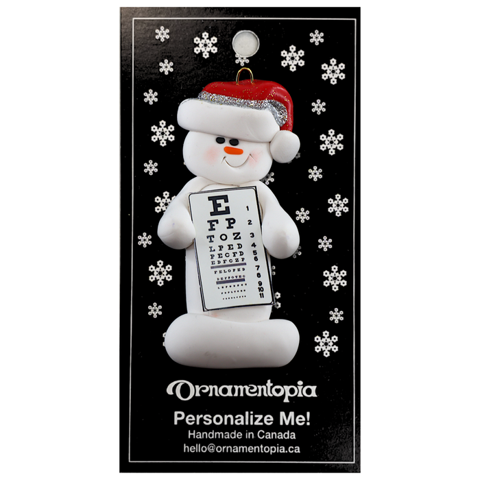 Ornament of white snowman in red hat holding an eye chart, on display backing card.