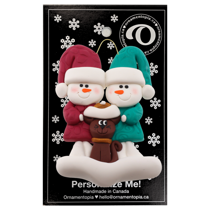 Ornament of snowman couple in red and green shirts and hats with a brown cat in Christmas hat, on a display backing card.